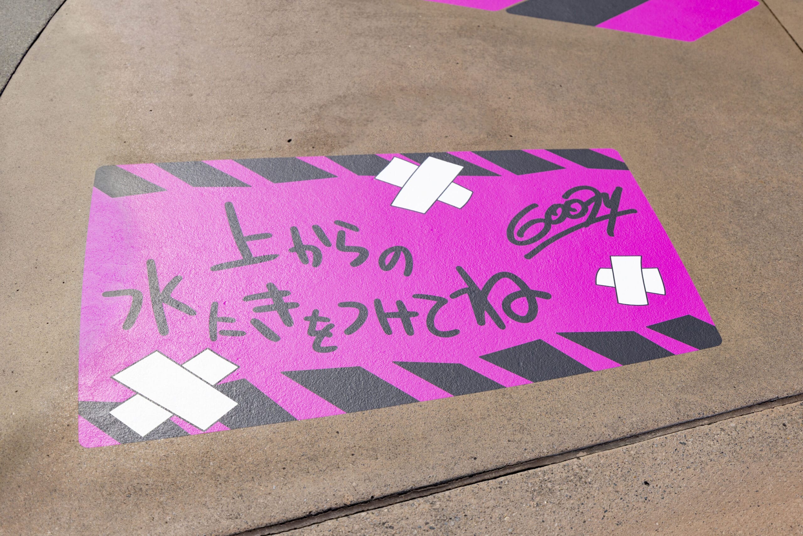 A pink taped sign with Japanese text on pavement, bordered with black stripes and white tape graphics.