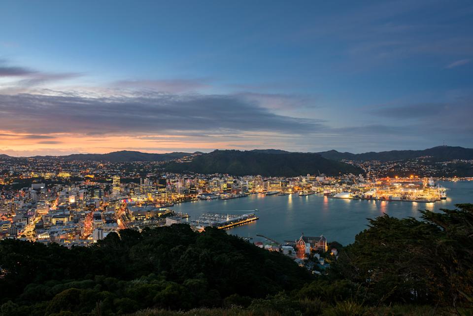 Cityscape of Wellington, New Zealand at dusk, featuring illuminated buildings, a calm harbor, and surrounding hills. Ideal for travel enthusiasts