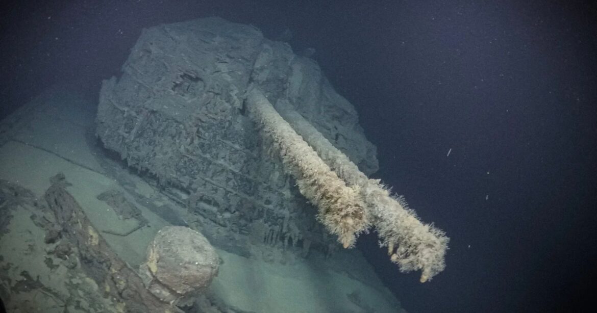 A sunken ship’s rusted, coral-encrusted gun turret lies deep underwater in dark, murky surroundings, showing significant decay and marine growth.