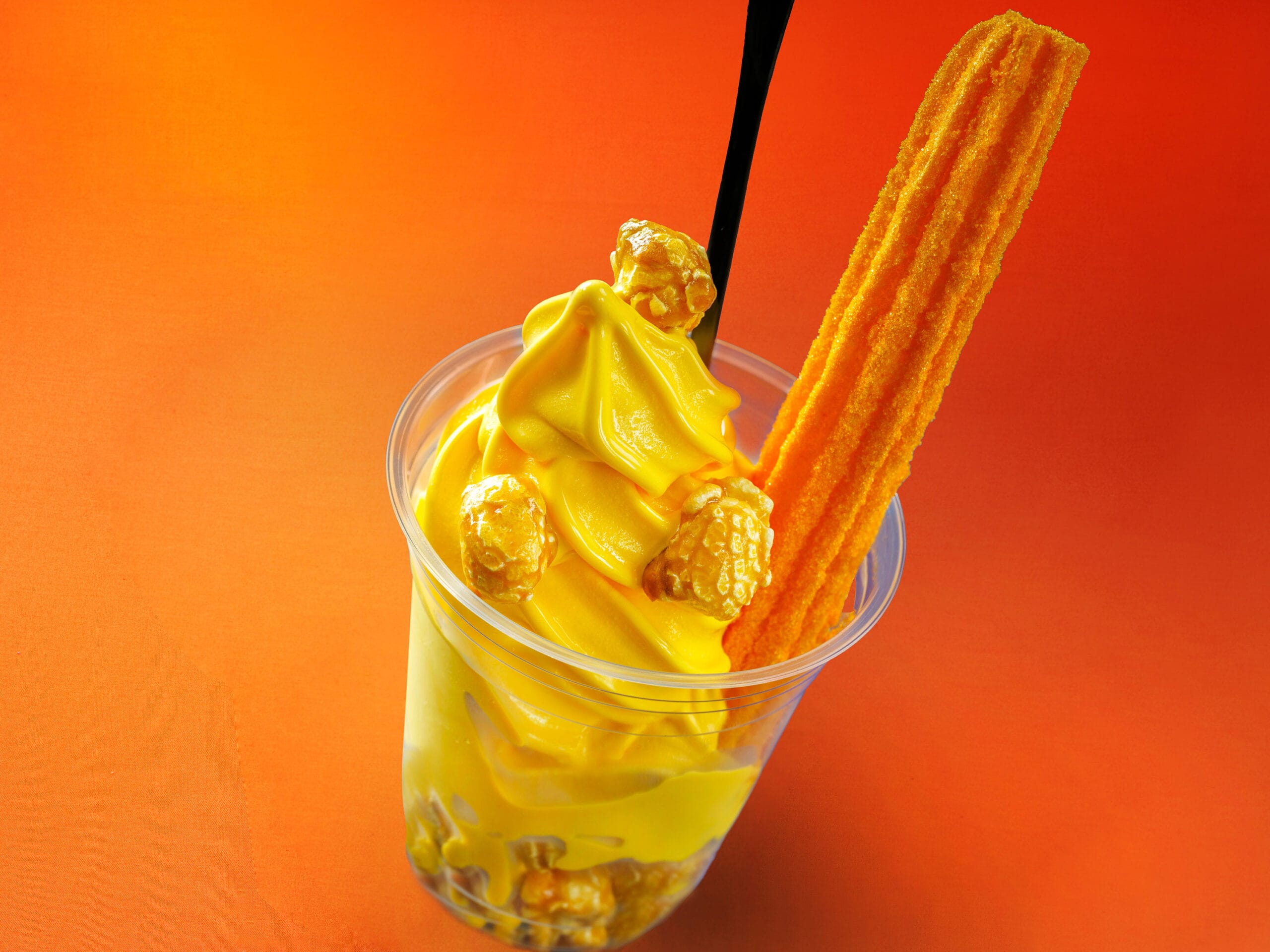Yellow soft serve at Tokyo Disney Resort, topped with caramel popcorn and a churro, against an orange Halloween backdrop.