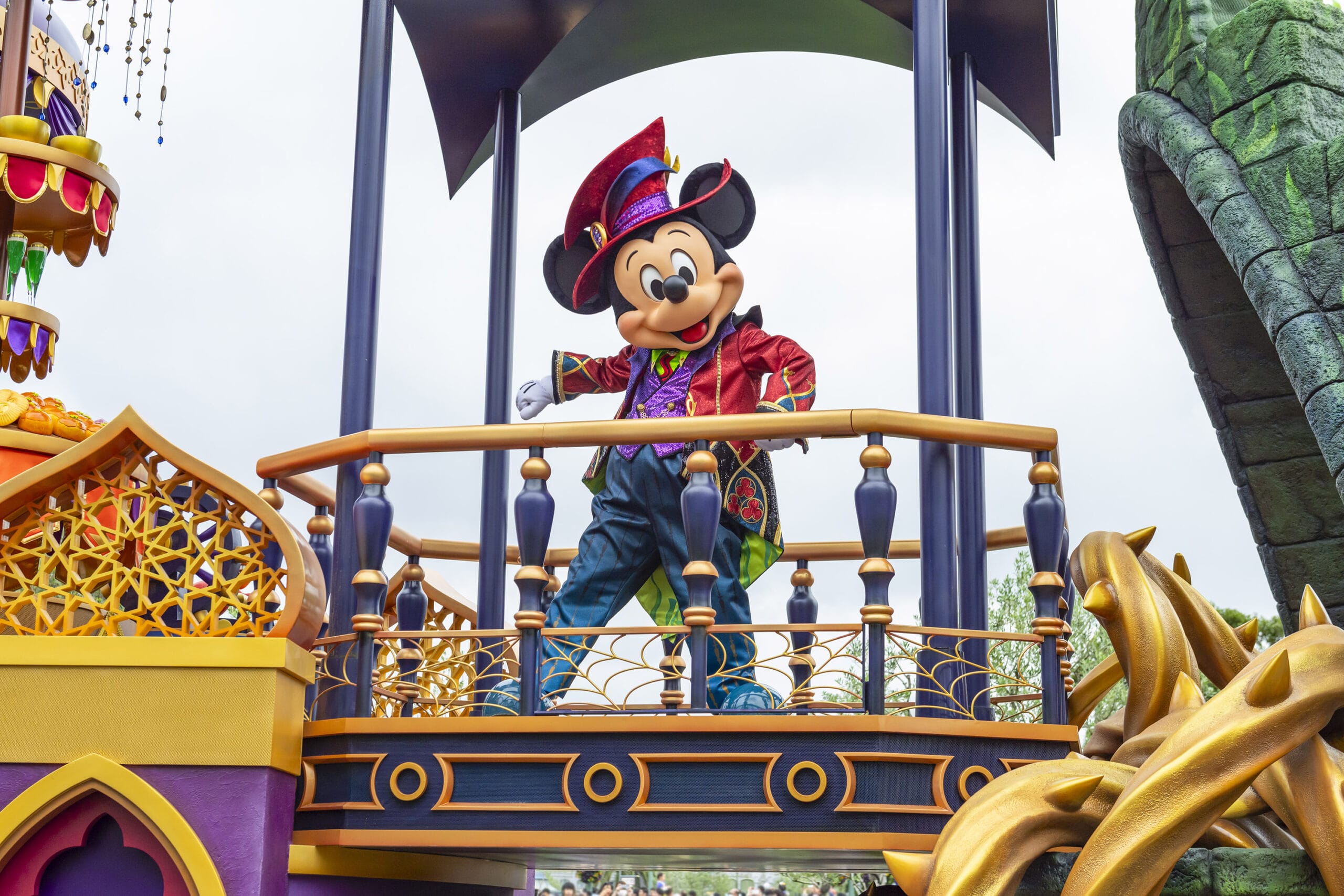 Mickey Mouse in a colorful Halloween outfit stands on a parade float, waving at Tokyo Disney Resort.