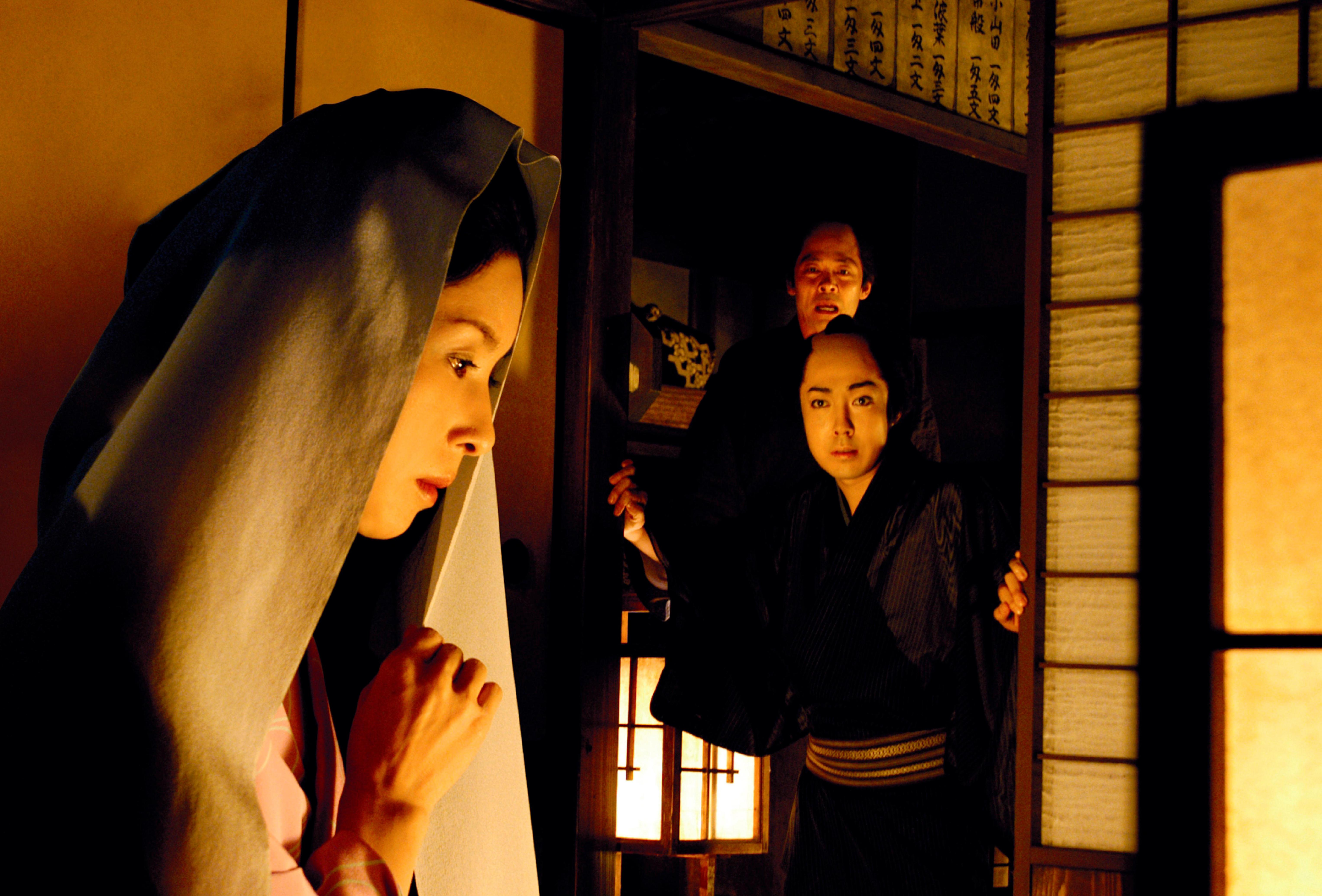 Still from a film showing a woman in a headscarf looking down, watched by two people in traditional Japanese clothing.