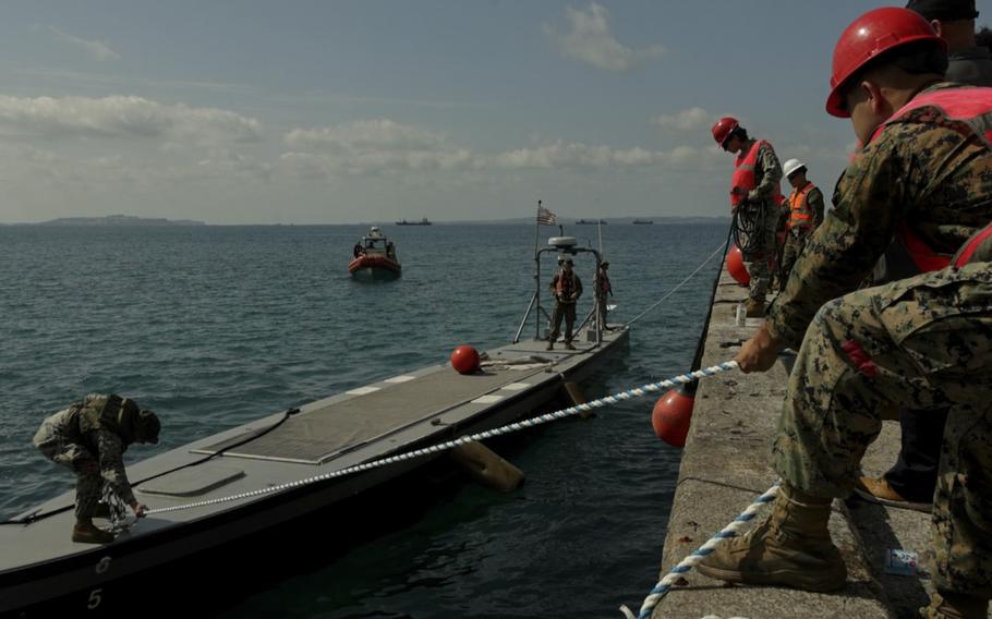 Marines wearing hard hats and safety vests pull a vessel towards a dock.
