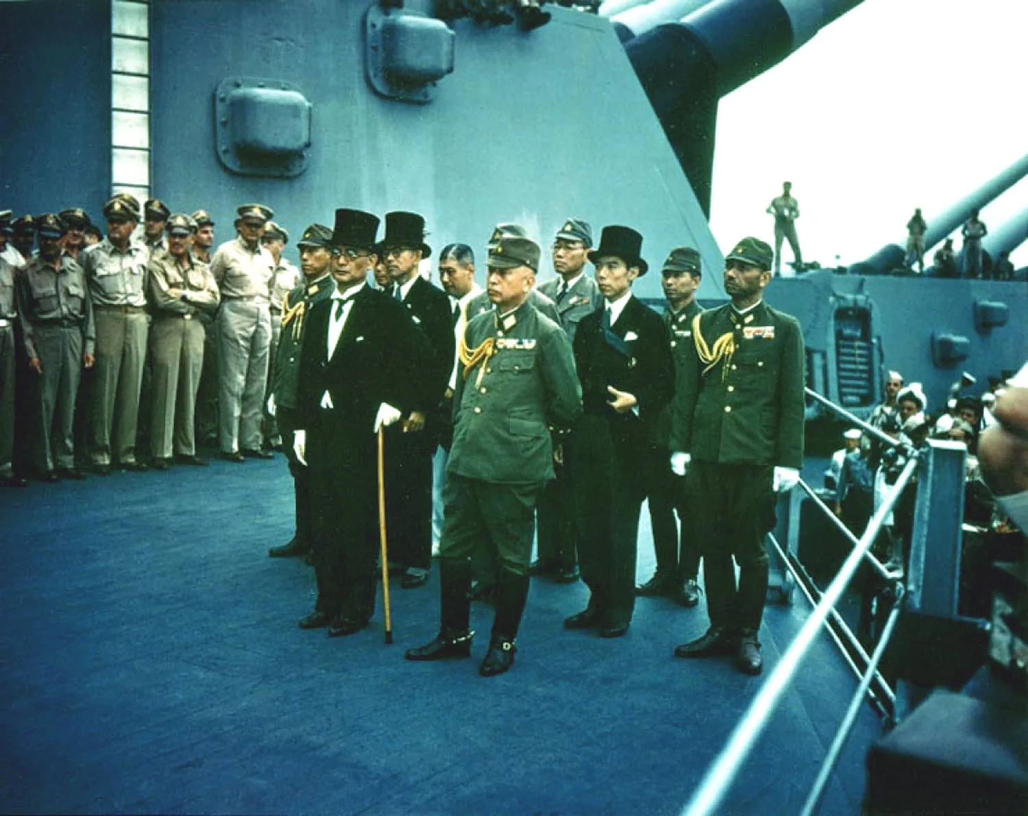 Officials in suits and top hats and others in military uniforms stand on the deck of a ship. U.S. men in uniform stand in the background.