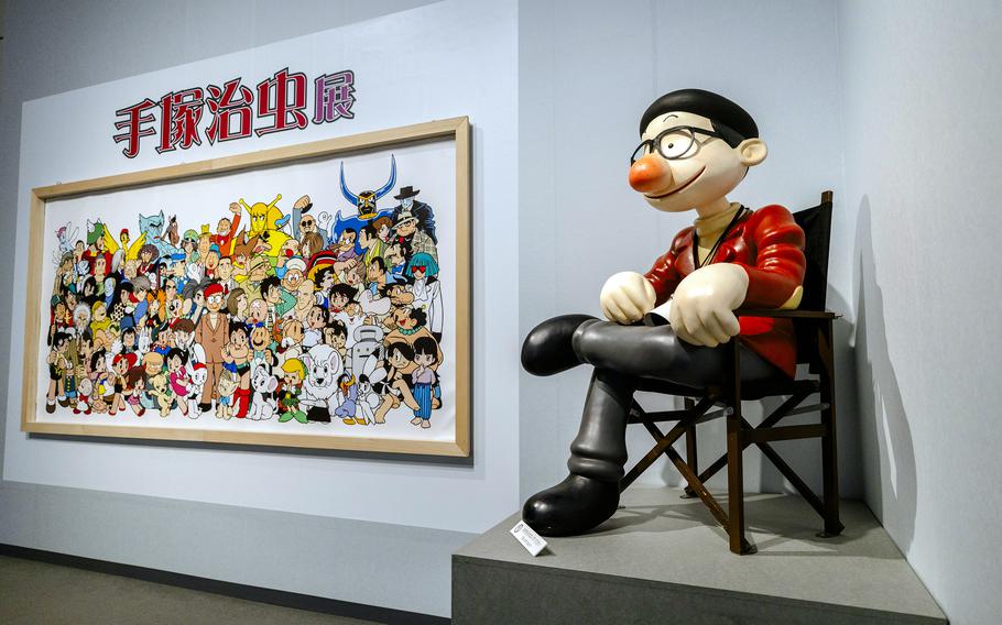 A large framed work depicts many manga-style cartoon characters, and a 3-D figure of a manga character is perched on a chair on a platform nearby.