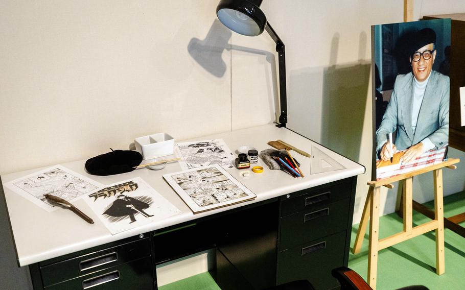 A desk displays artists’ implements and drawings, and an easel nearby shows a man’s photo.