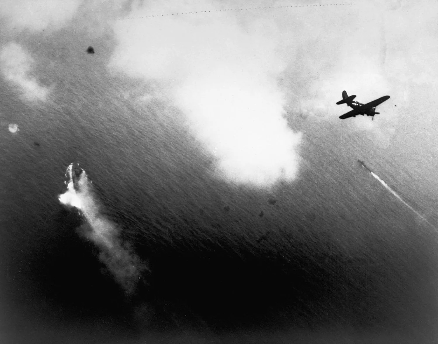 A smoking battleship is seen from above as planes fly over.