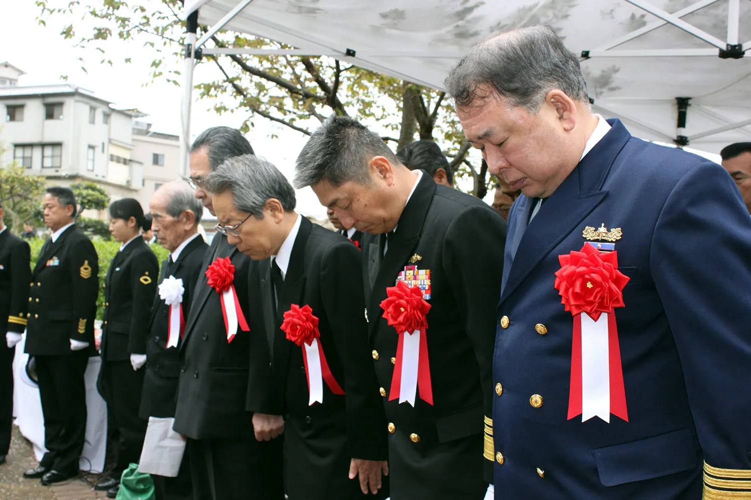 Men in uniforms wearing large ribbon rosettes pinned to their chests bow their heads.