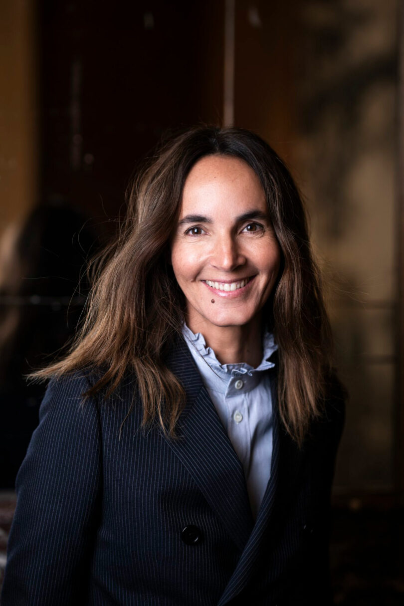 Dorothée Boissier, with long brown hair, wearing a pinstripe blazer and light blue blouse, smiles at the camera in a dimly lit indoor setting.