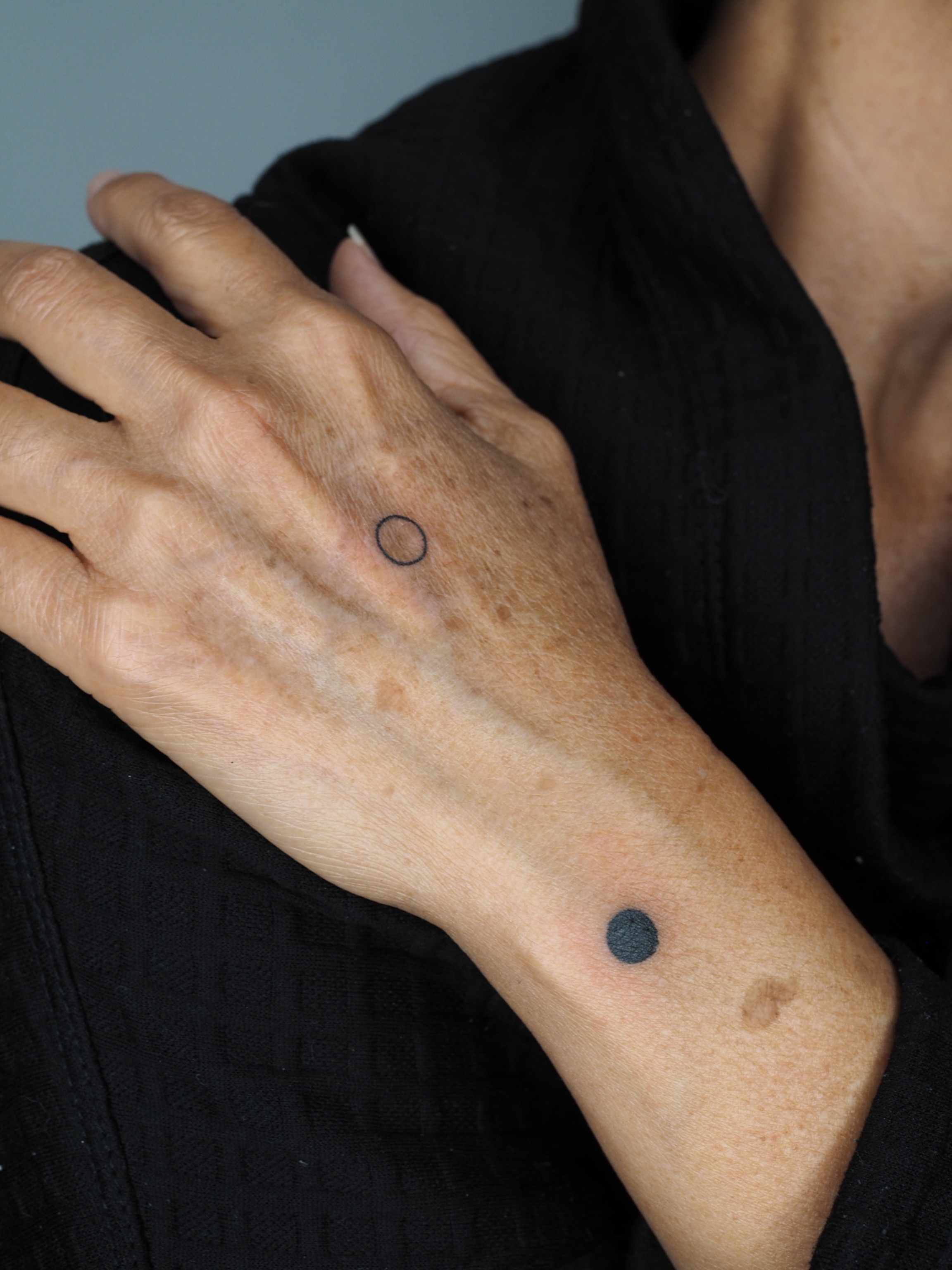 A person's left hand, tattooed with a black circle on the back of their wrist and the outline of circle on the back of their hand.