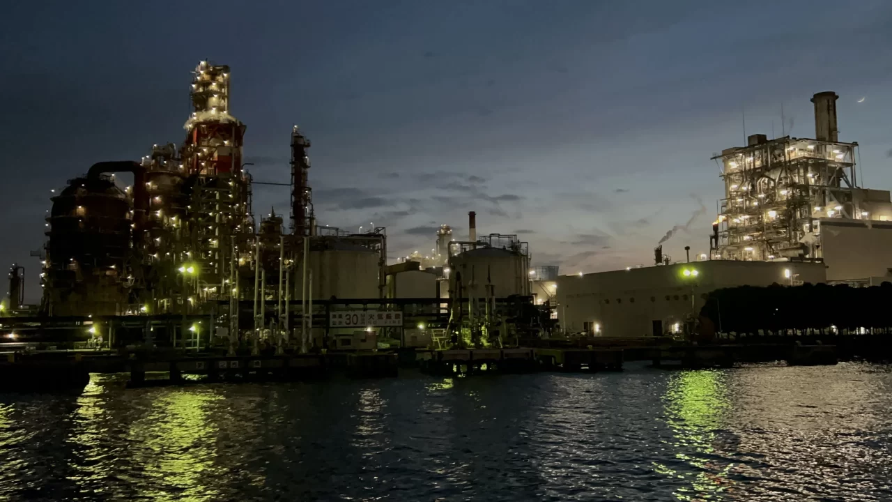 Photo of the Keihin Industrial Zone outside the port of Yokohama. Factories glow with neon lights reflecting on the water. 