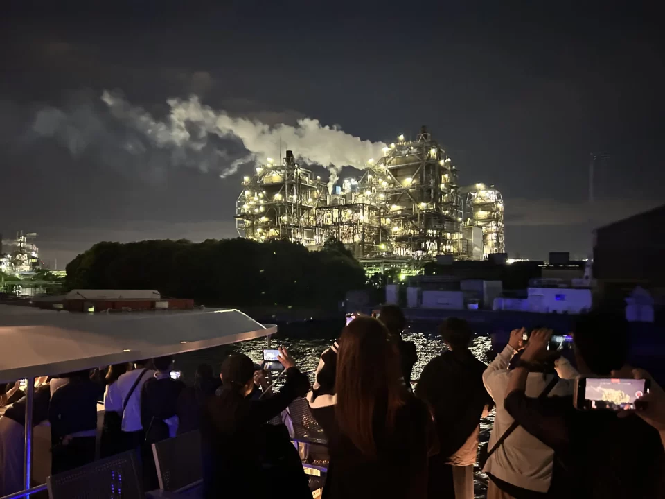Photo of the Keihin Industrial Zone outside the port of Yokohama. A factory flashing with bright lights can be seen, smoke pouring from its top. 