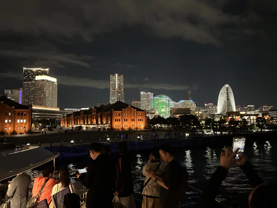 Photo of Minato Mirai taken at night. Yokohama's buildings glow with bright lights, and the Red Brick Warehouse buildings can be seen across the water. 