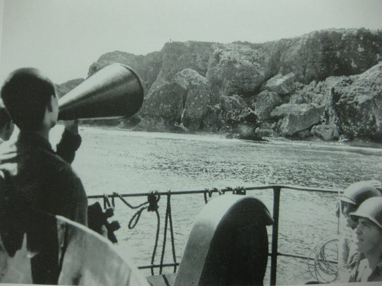 Archival photograph of a Japanese POW shouting through a loudhailer from a ship to an island.