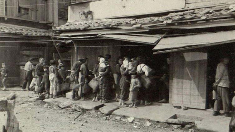 A sepia-coloured image of a group of Japanese people queueing on a street for food.