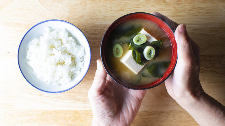 Hands holding bowl of miso soup next to bowl of rice