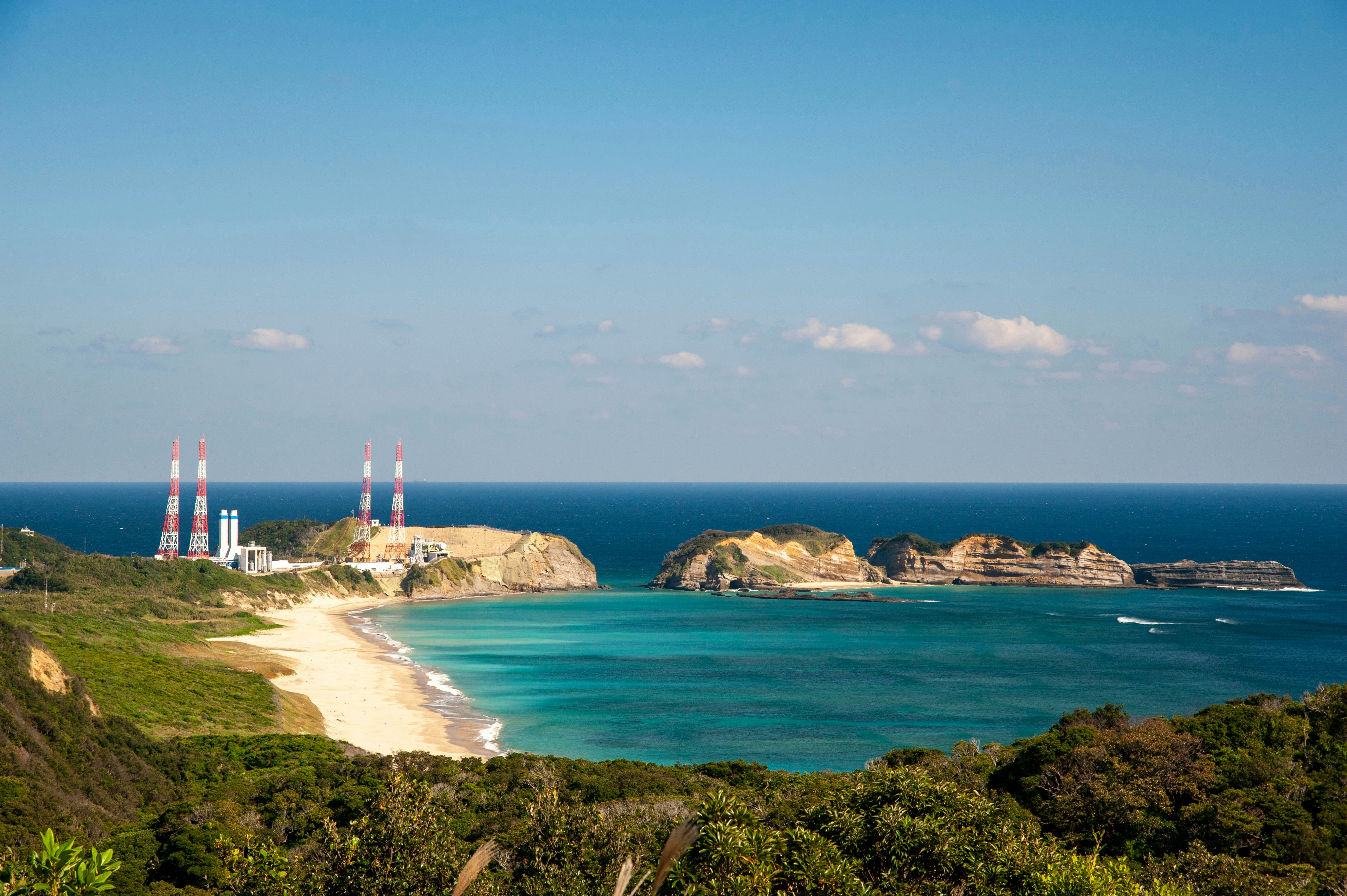 A sandy bay lapped by turquoise waters. A rocket launching pad stands at one end of the promontory.