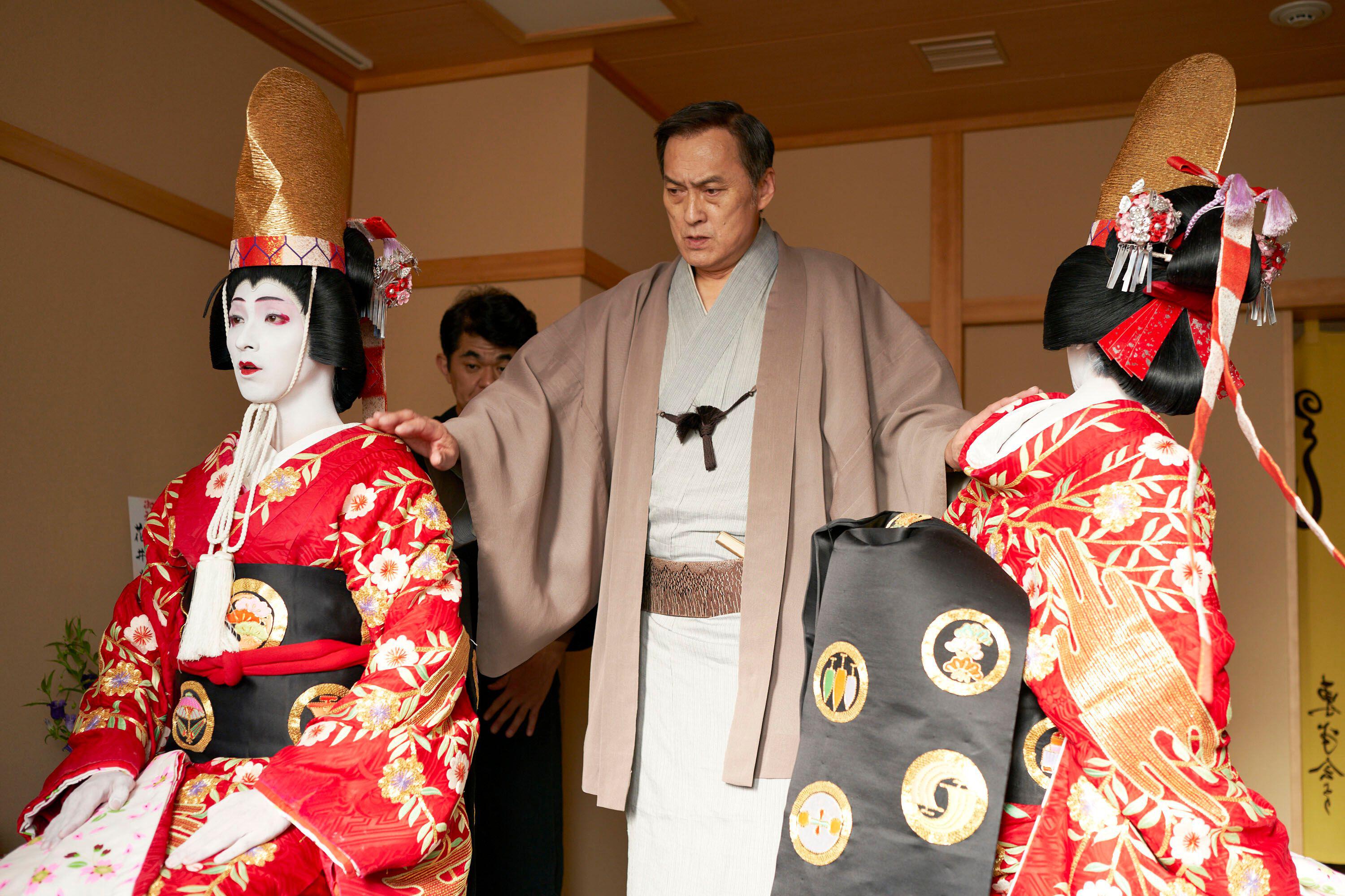 Film still of a man in traditional Japanese clothing assisting two women in elaborate kimonos.