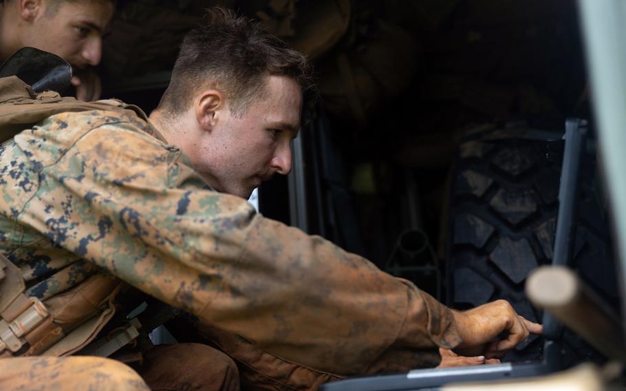 A military service member in a camouflage uniform operates equipment inside a military vehicle with another service member visible in the background.