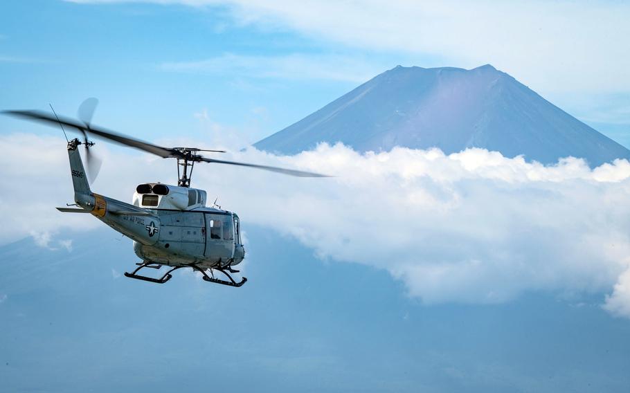 A U.S. Air Force helicopter flies against a backdrop of blue sky with white clouds and a large mountain in the distance. 