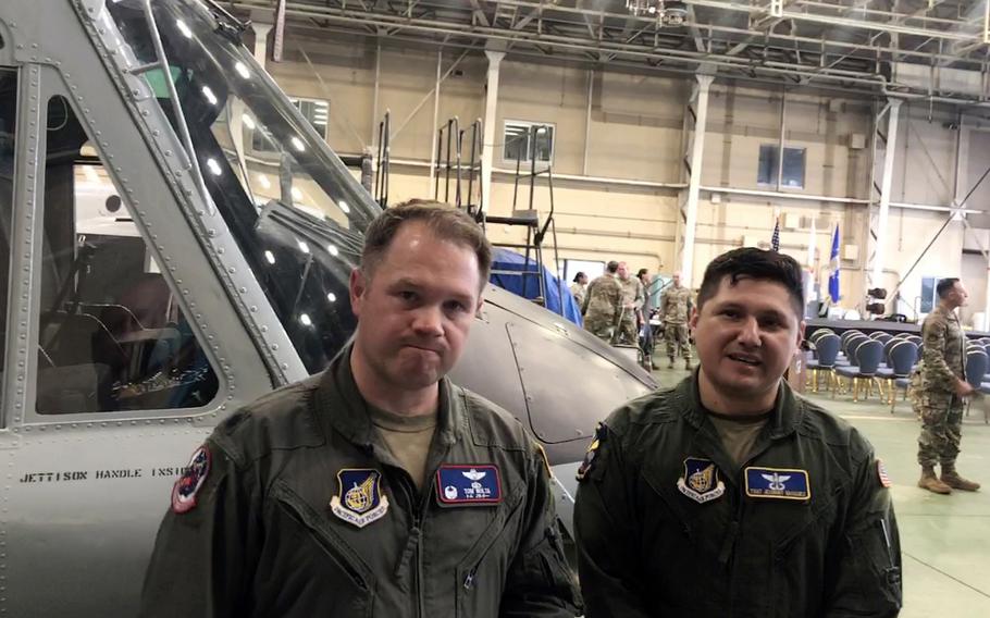 Two U.S. Air Force personnel in green flight suits pose for a photo in front of a military helicopter inside a hangar.