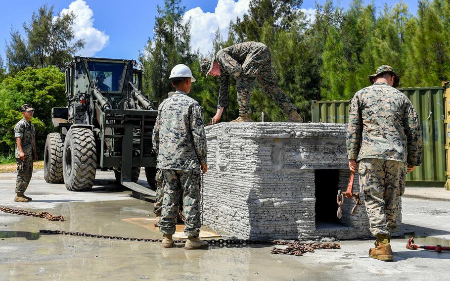 Two Marines in camouflage uniforms stand next to a concrete structure, while another Marine in camouflage leans over while standing on top of the structure and a fourth Marine sits in the driver’s seat of a heavy lifter in the background.