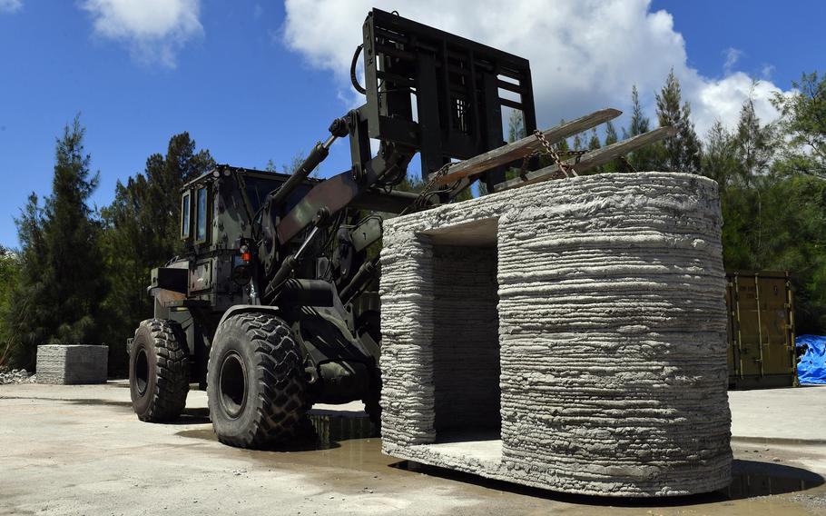 A concrete structure with a doorway-like hole in the side sits next to a heavy lifter with a camouflage paint job.