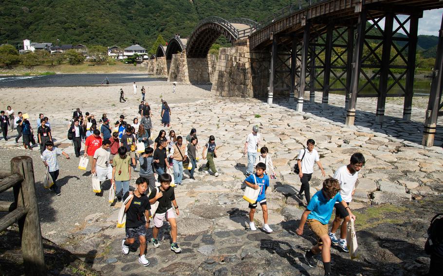 Dozens of students walk up a manmade rocky shoreline with Kintaikyo Bridge in the background.