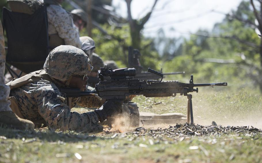 A Marine in digital woodland camouflage uniform fires a machine gun mounted on a bipod during outdoor field training. Dust kicks up around the weapon as other Marines are visible in the background among trees and vegetation.