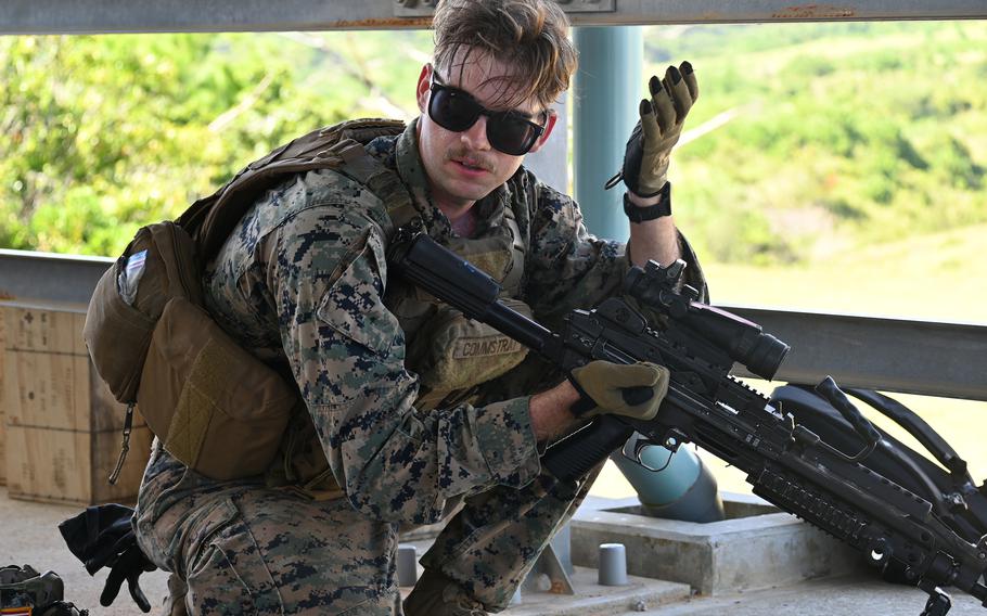 
A Marine wearing sunglasses and digital camouflage uniform signals with his raised hand while holding an assault rifle during training exercises. He is positioned behind concrete barriers with green foliage visible in the background.