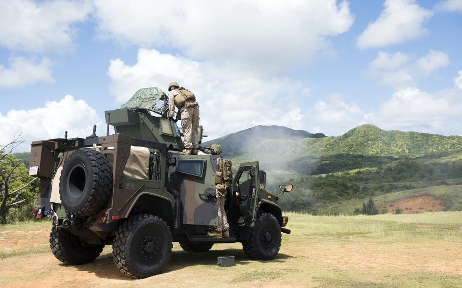 
Military personnel work on a camouflaged tactical vehicle in a field setting. One service member stands on top of the vehicle while another is positioned nearby, with lush green mountains visible in the background under a cloudy sky.