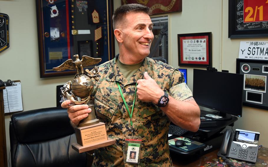 A Marine in camouflage uniform holds his left fist to his chest and holds a trophy in his right hand while standing in an office with framed memorabilia on the walls behind him.