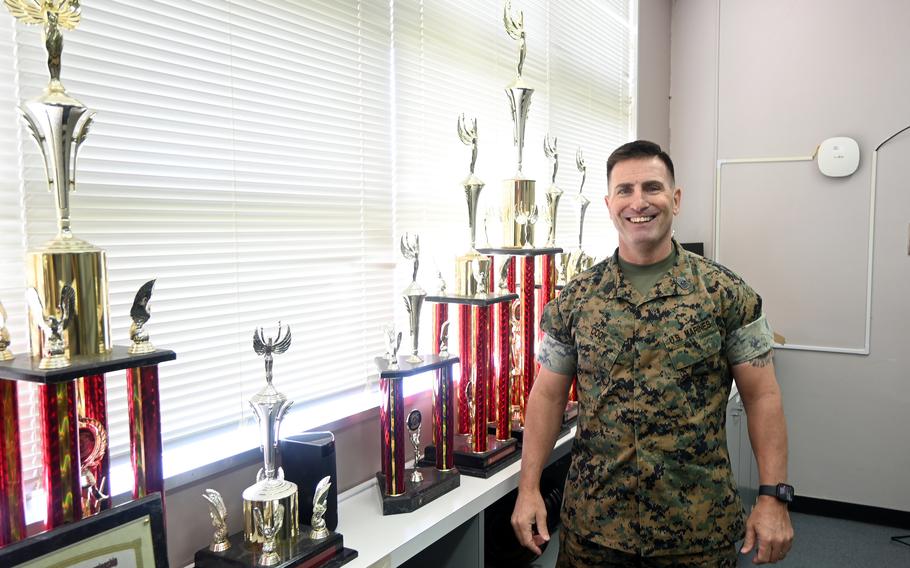 A Marine in camouflage uniform smiles while posing next to a shelf with several trophies in front of a window with white blinds.