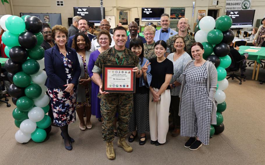 A Marine in camouflage uniform smiles and holds up a framed certificate while posing with several teachers next to strands of balloons in a school office.