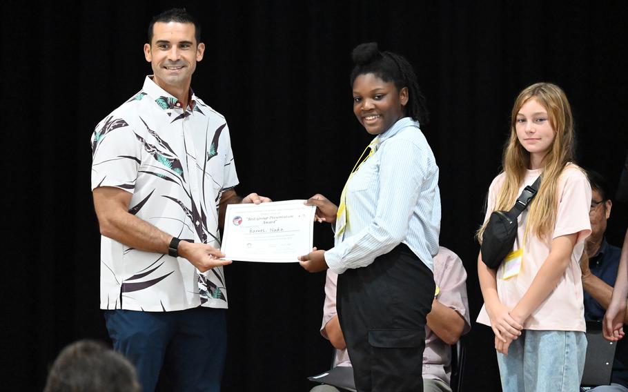 A middle school girl poses with an airman who hands her an award for best presentation.