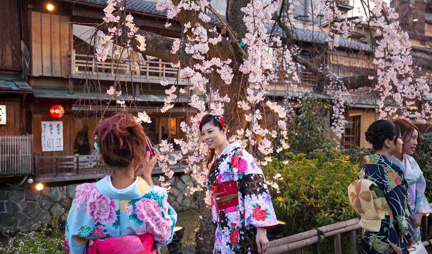 Young woman in kimono smiles at the camera with cherry blossoms behind her