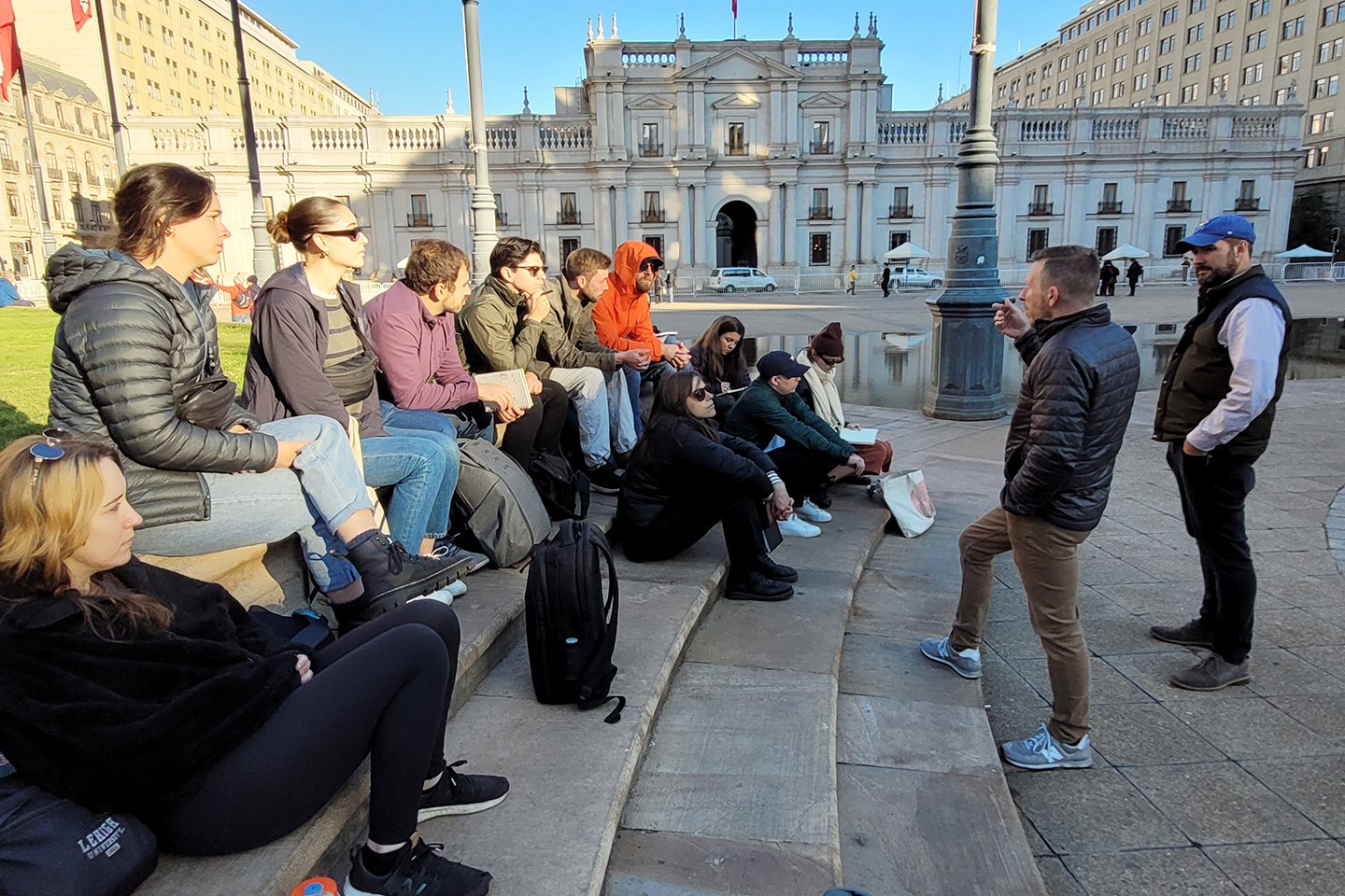 Ken Schroppel and Andy Rutz speaking to a group of students sitting on the steps of a plaza. 