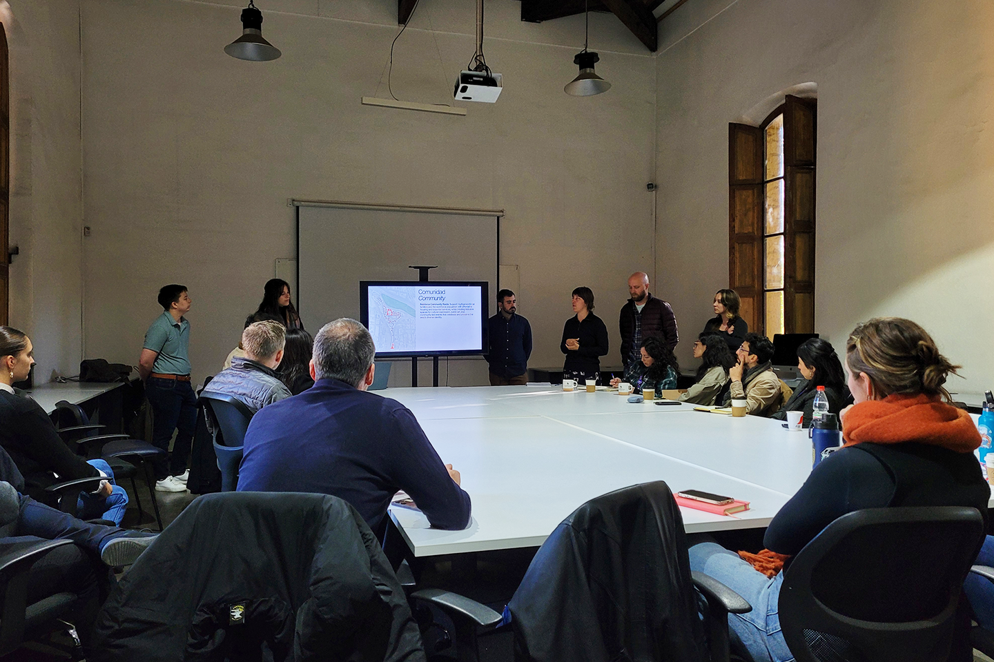 A room full of people listening to a presentation by six students at the front of a classroom with a screen centered between the student presenters. 