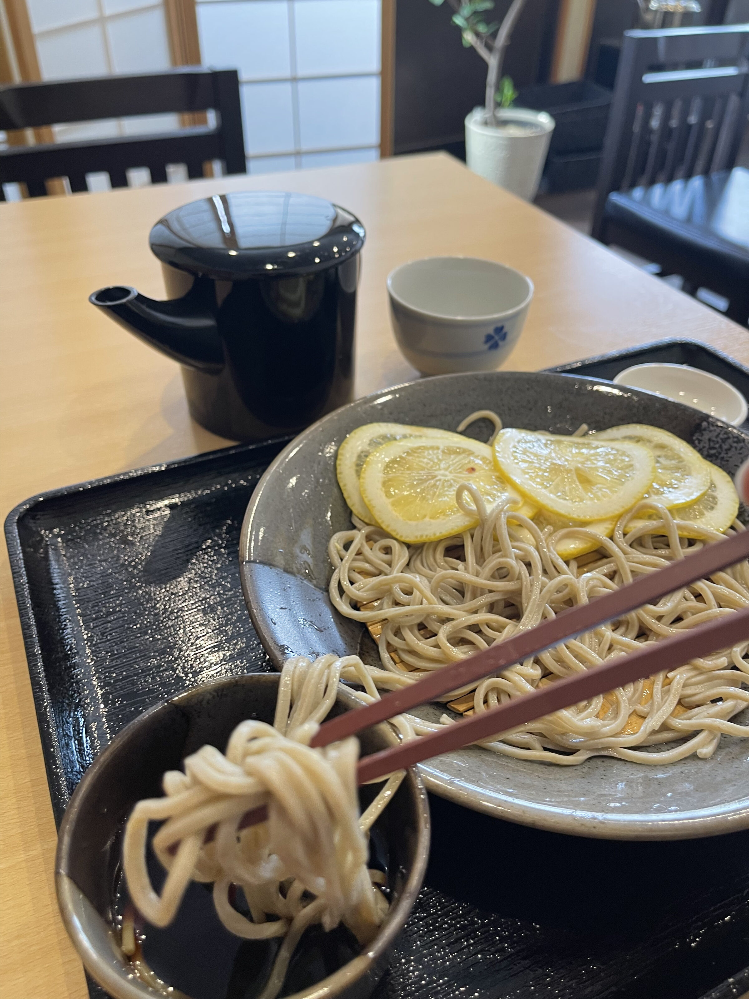 A bowl of cold soba noodles, with sliced lemon on top. Chopsticks dipping some of the noodles into a soy sauce cup are in the foreground