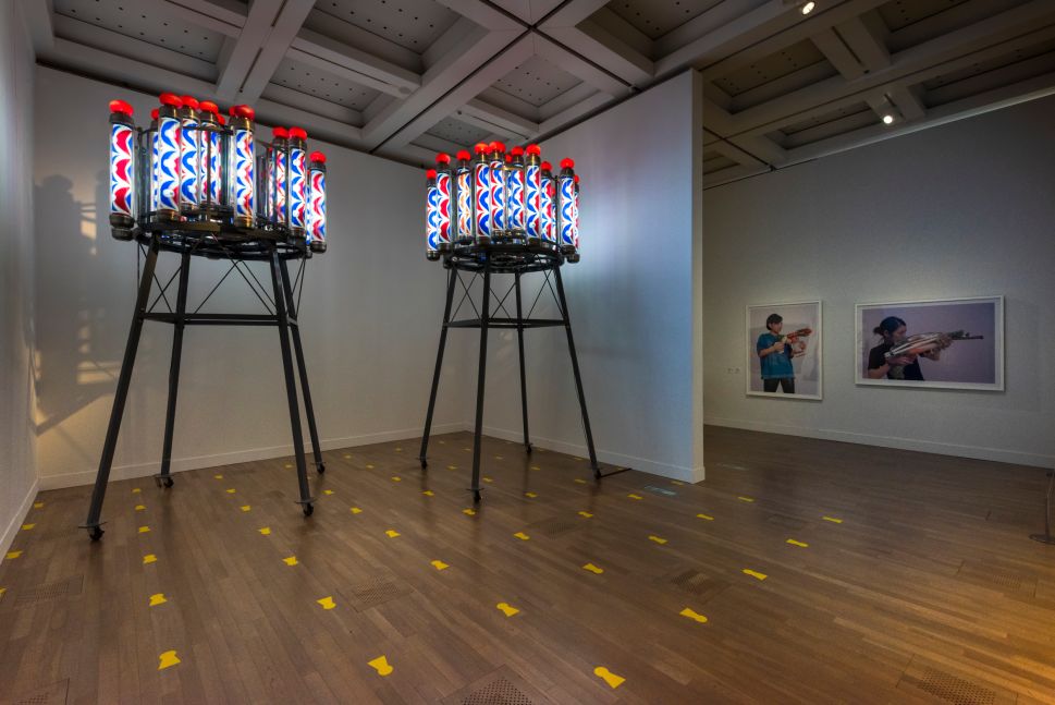 A gallery installation featuring two tall black metal structures topped with illuminated cylindrical tubes patterned in red, blue, and white, with yellow tape markers on the floor and large photographs of people holding makeshift weapons displayed on the adjacent wall.