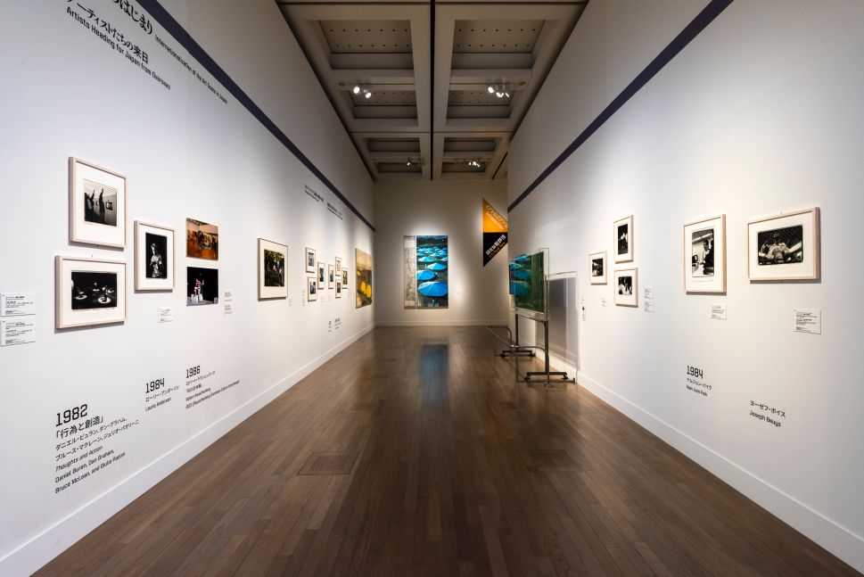 A long gallery corridor with framed photographs and artworks arranged chronologically on both walls, featuring black-and-white and color images, wall text with years such as 1982, 1984, and 1986, and a large photograph of blue umbrellas displayed at the far end.