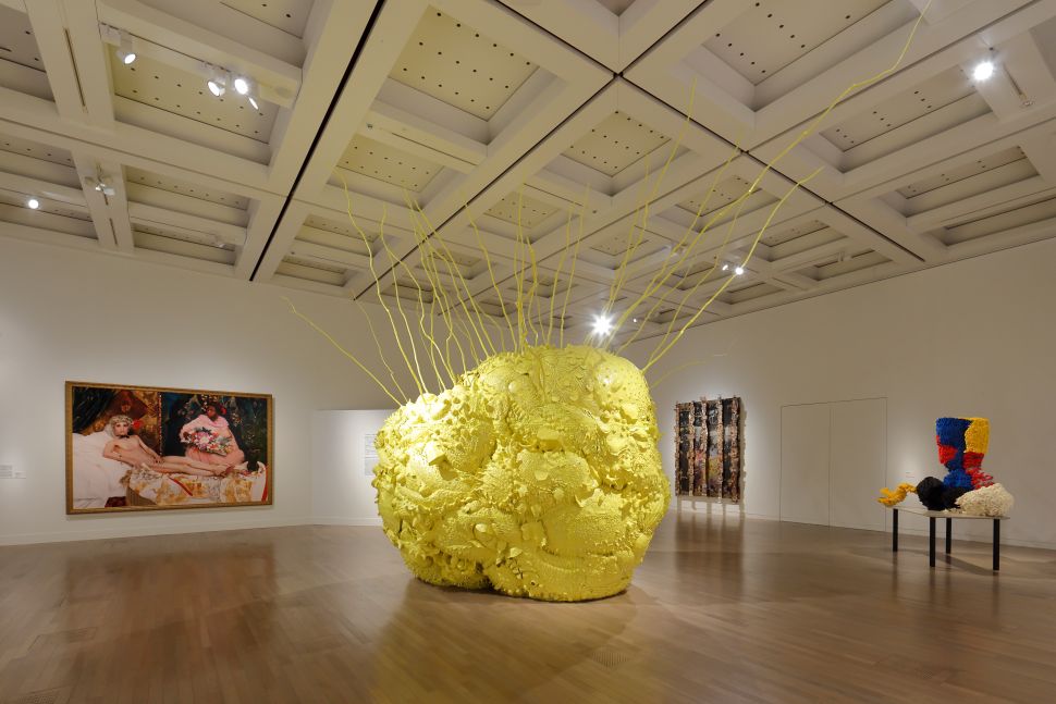 A bright yellow, bulbous sculpture with spindly branches extending upward, displayed alongside colorful textile works and a figurative painting.