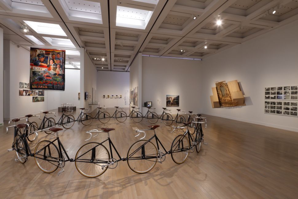 A gallery installation featuring a circle of bicycles connected wheel to wheel, with large-scale photos and sculptural works on surrounding walls.