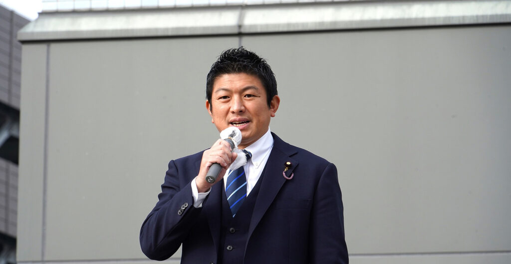 Sohei Kamiya wearing a dark blue suit and tie talking into a microphone.