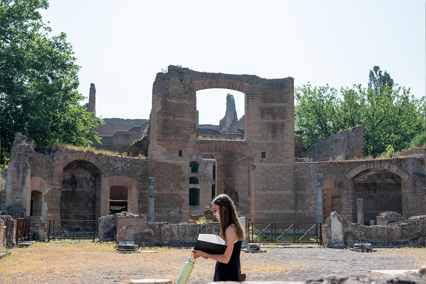 A student walks past an ancient Roman structure while holding her sketchbook and a bottle of water.