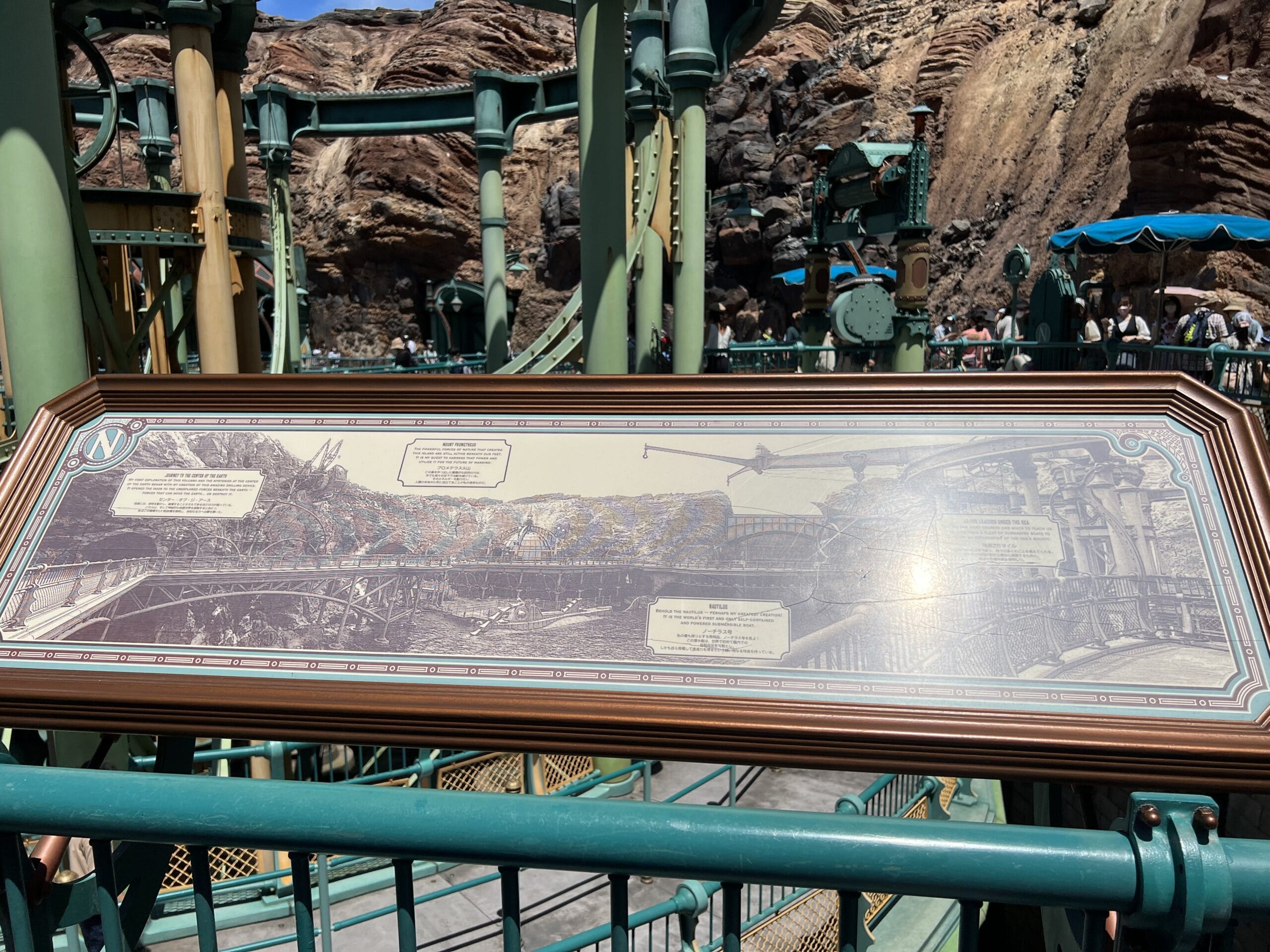 Framed illustrated map signage with labels, set outdoors near Mount Prometheus among green railings and rocky cliffs.