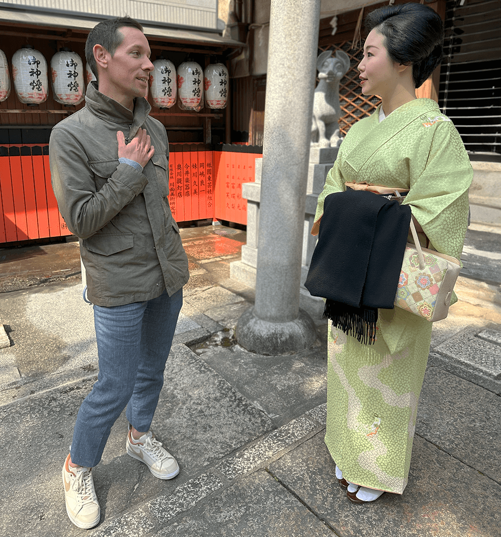 Tyler is speaking to Tomitsuyu, a geisha from Kyoto. They are standing outside, on a traditional street in Gion.