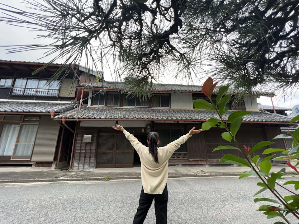 A woman standing in front of a traditional farmhouse in the Japanese countryside.