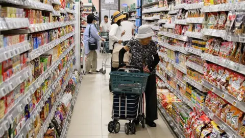 Getty Images An elderly woman wearing a white hat and dark clothing browses the crowded shelves at a Japanese supermarket, as she pushes a shopping trolley.