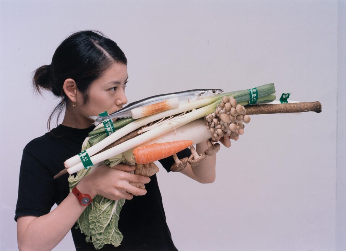 Must-See Tokyo Exhibitions: Delaunay, Katz, Fujimoto and More A woman in a black shirt holds a playful assemblage of vegetables and a fish arranged like a rifle, aiming it as if it were a weapon; the composition includes leeks, carrots, mushrooms, cabbage, and a whole fish.